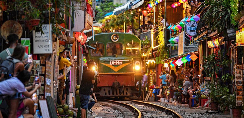 A train passing through Hanoi Train Street near Long Bien Station with people sitting at cafes